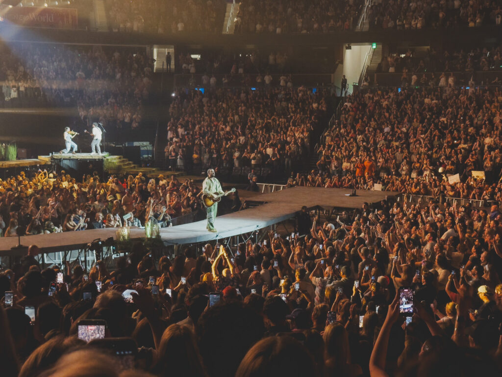 Wide arena shot, Noah Kahan on the thrust stage with acoustic guitar, Kia Center Orlando