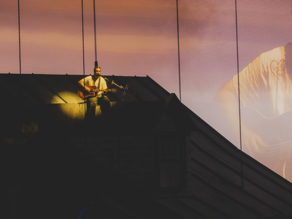 Noah Kahan performing We Go Way Back from the roof of the house set at sunset projection