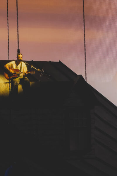 Noah Kahan performing We Go Way Back from the roof of the house set at sunset projection