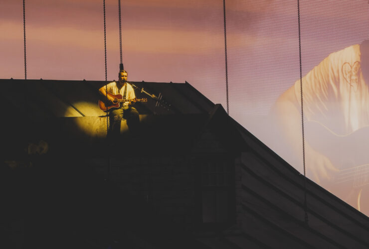 Noah Kahan performing We Go Way Back from the roof of the house set at sunset projection