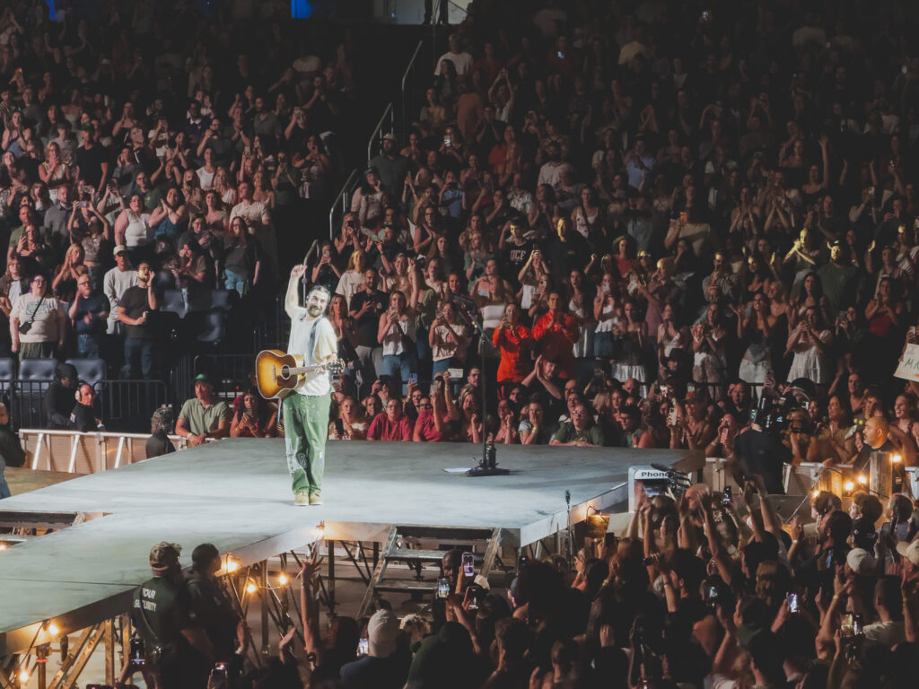 Noah Kahan raising his hand to the crowd on the thrust stage, Kia Center